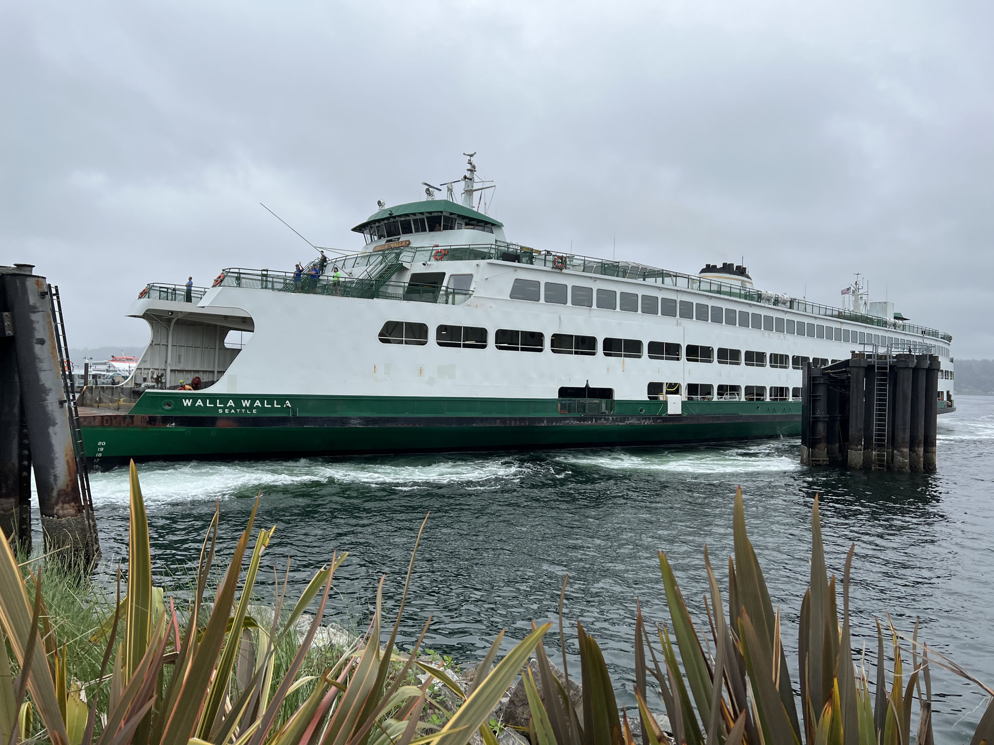 The Walla Walla ferry docked near a pier with green plants in the foreground and cloudy sky overhead.