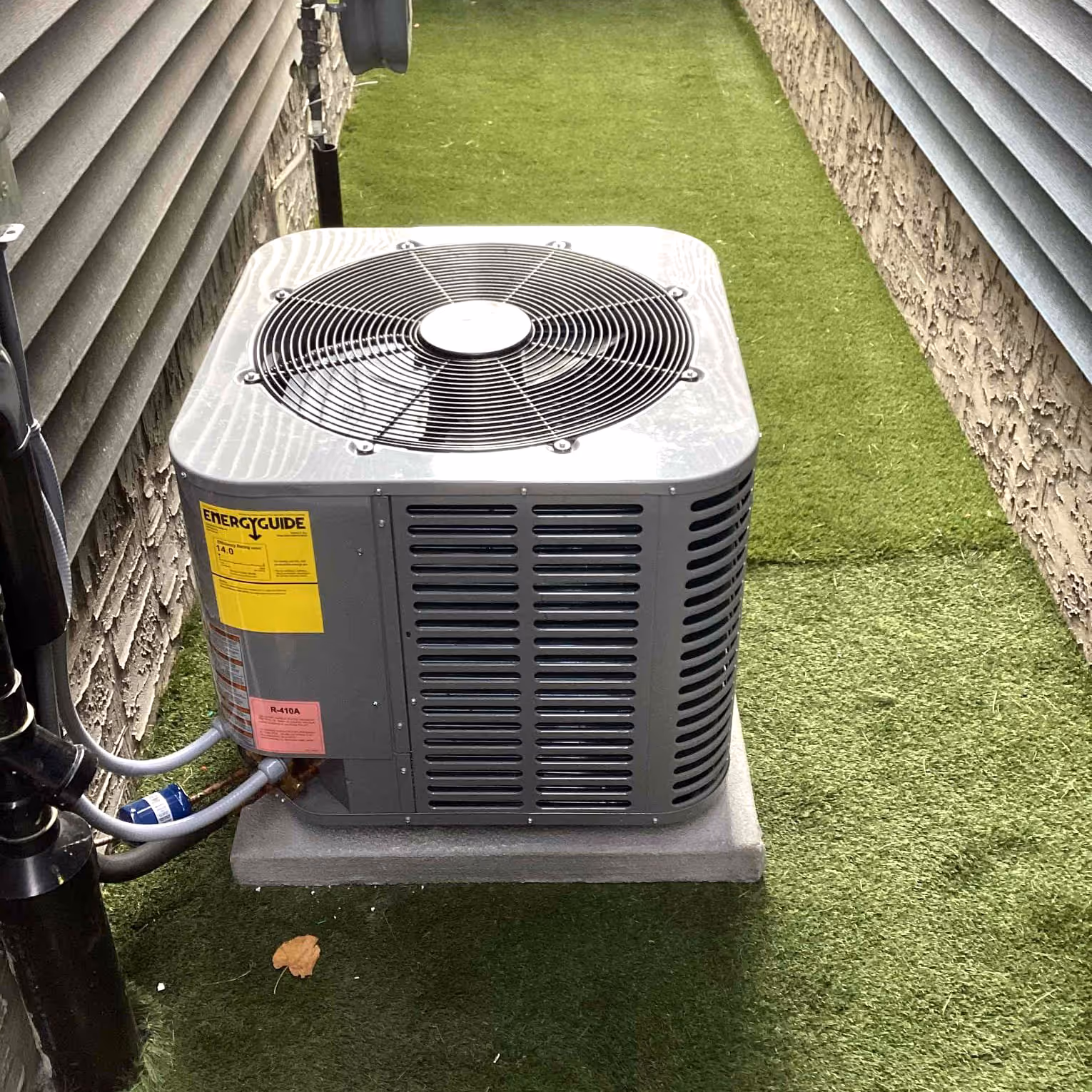 Outdoor gray air conditioning unit on artificial grass between house walls.