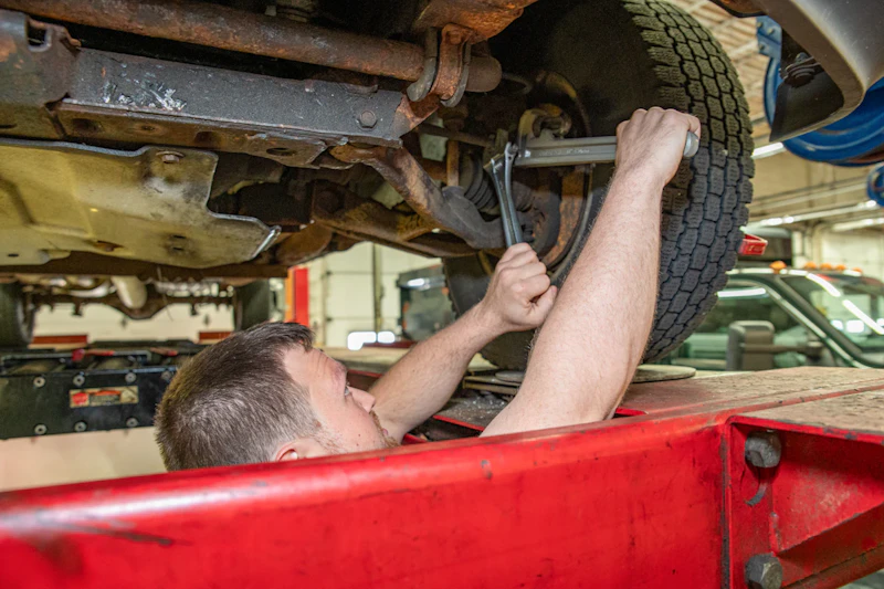 Heavy-duty truck technician performing axle repair underneath a lifted truck, using a wrench to service the suspension and axle components in a professional repair shop.