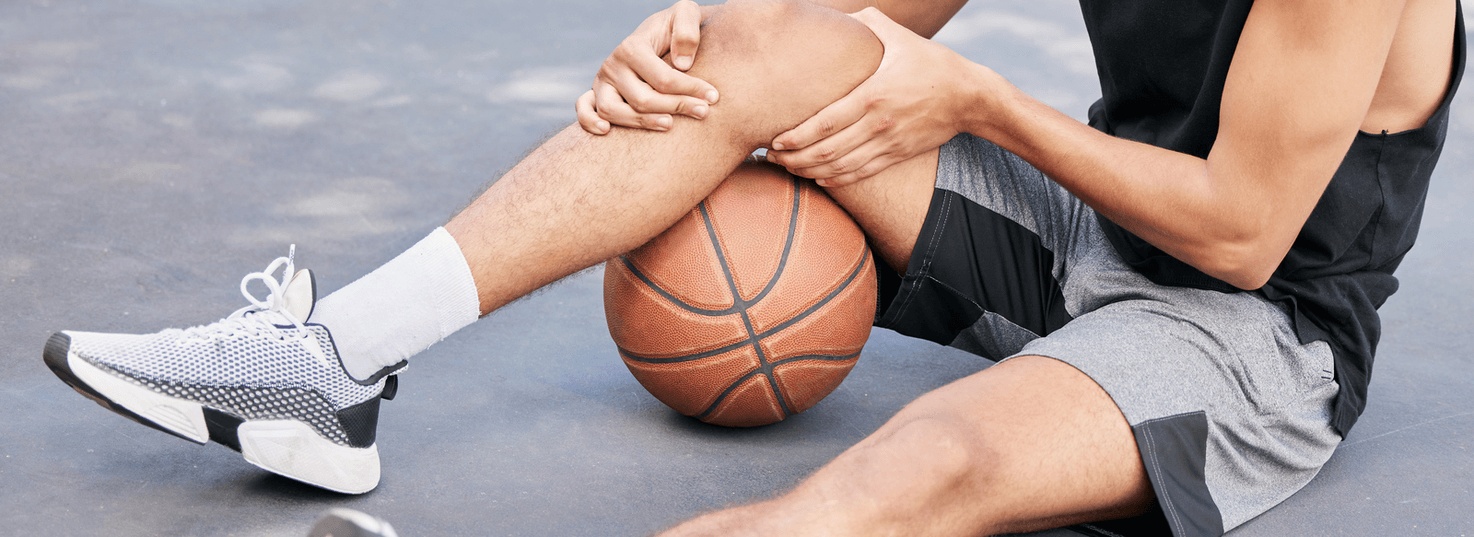 Athlete is sitting on an outdoor basketball court holding his knee, showing signs of a knee injury and seeking care from the best orthopedic doctor in Encinitas.