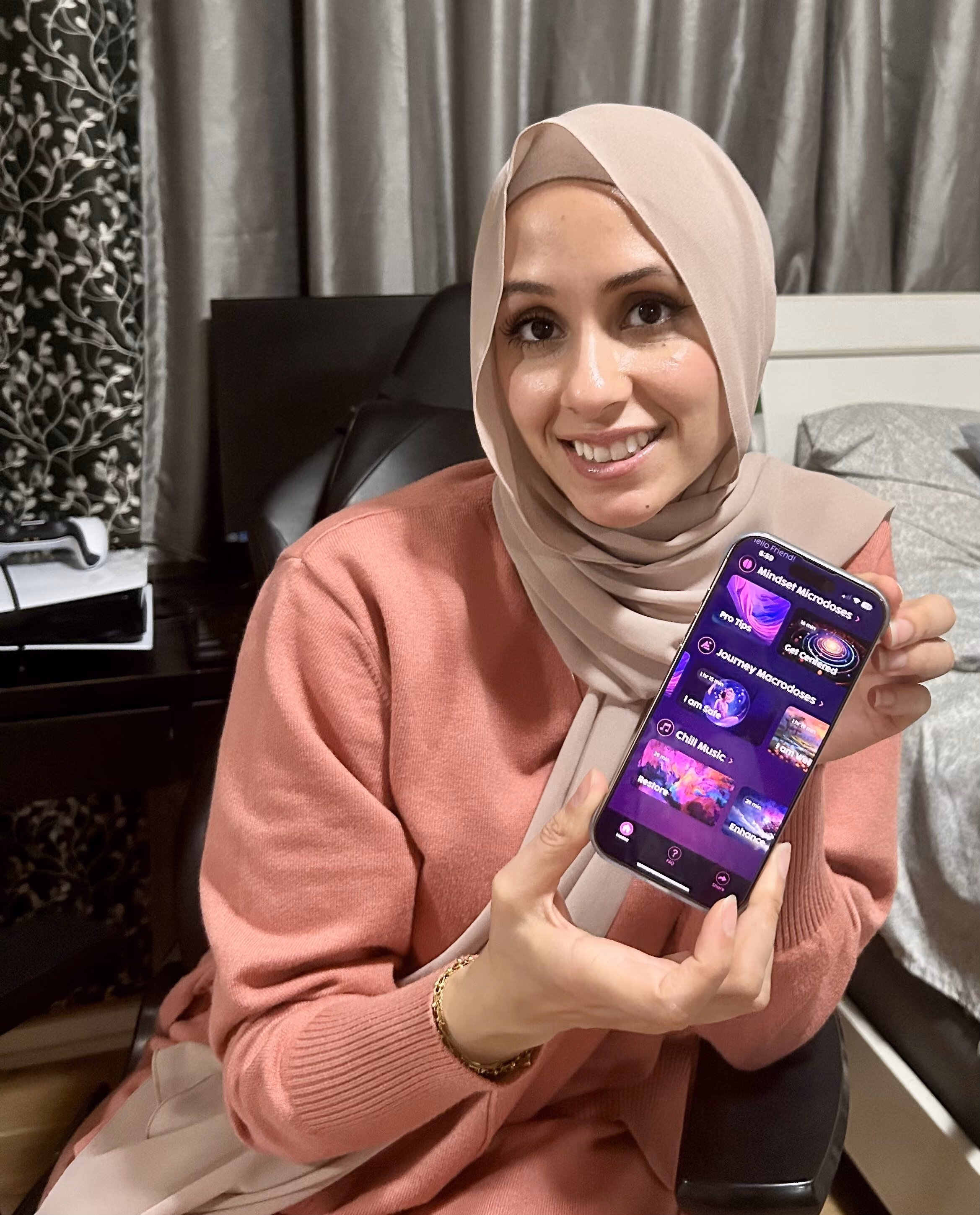 Smiling woman wearing a beige hijab and pink sweater showing a smartphone with a purple-themed Limitless mindset companion app interface.