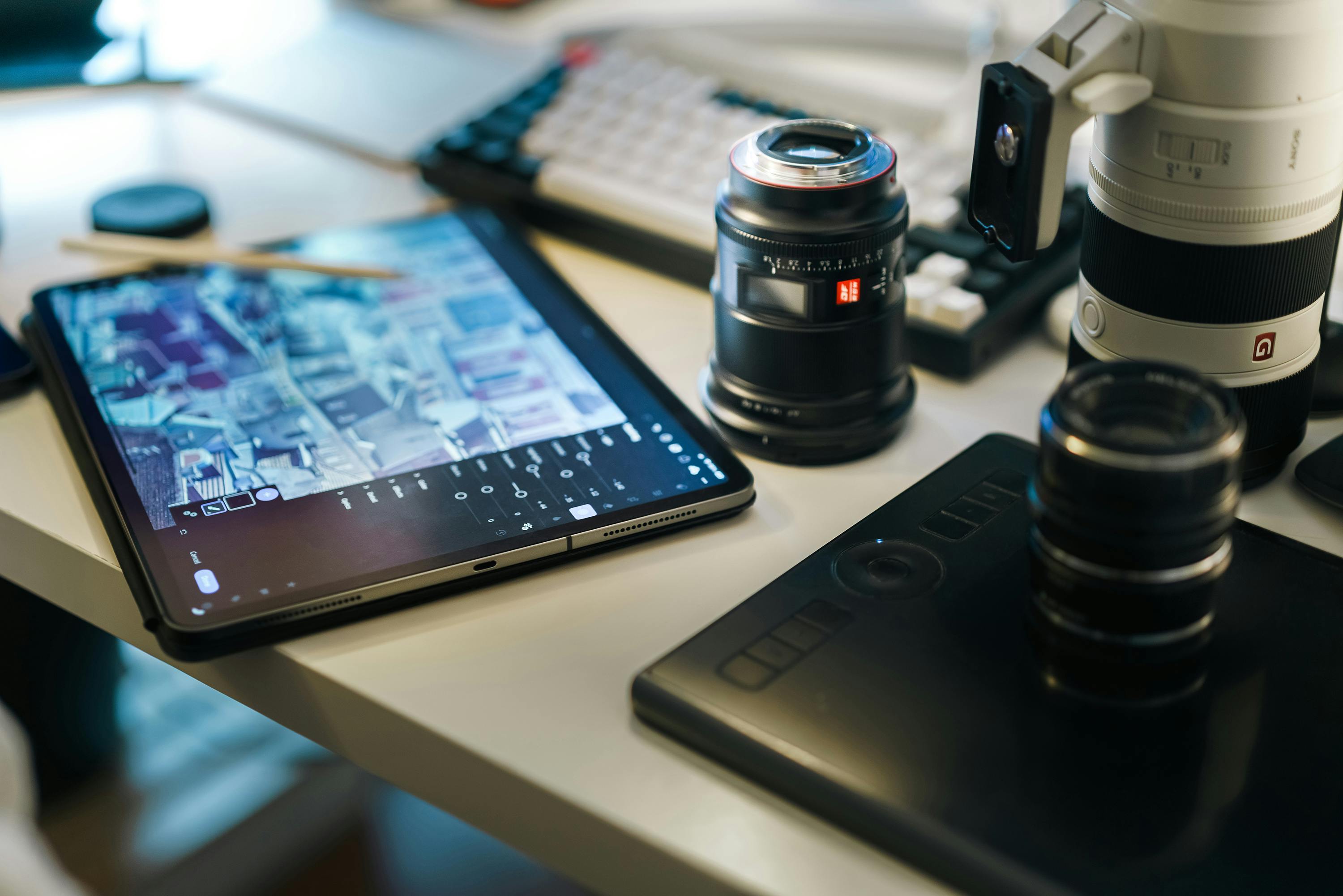 A photography workspace with a tablet displaying editing software, multiple camera lenses, and a graphics tablet on a cluttered desk, conveying creativity.