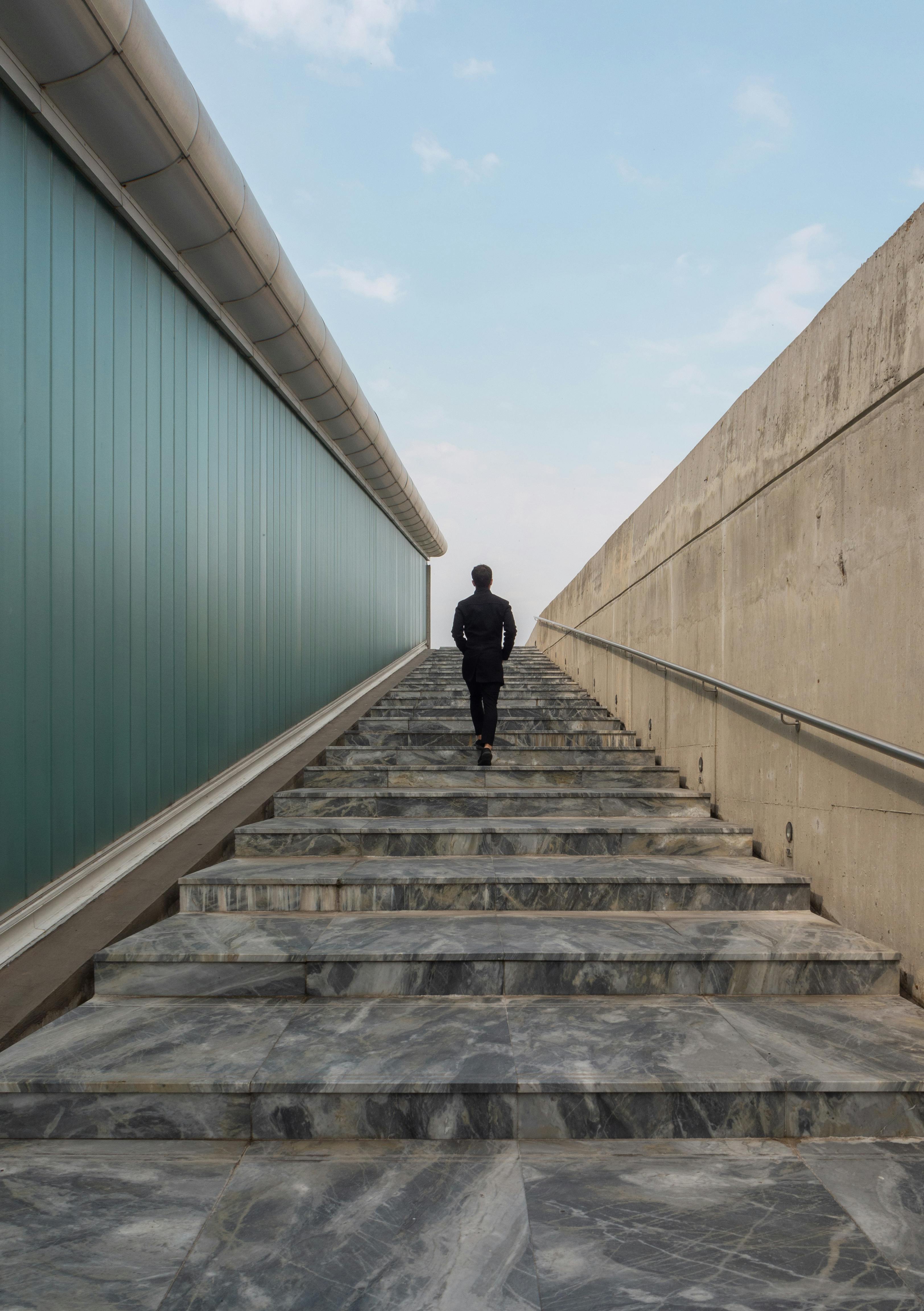 A person in dark clothing ascends a long, marble staircase flanked by a glass wall and concrete barrier, with a clear sky above, evoking solitude.