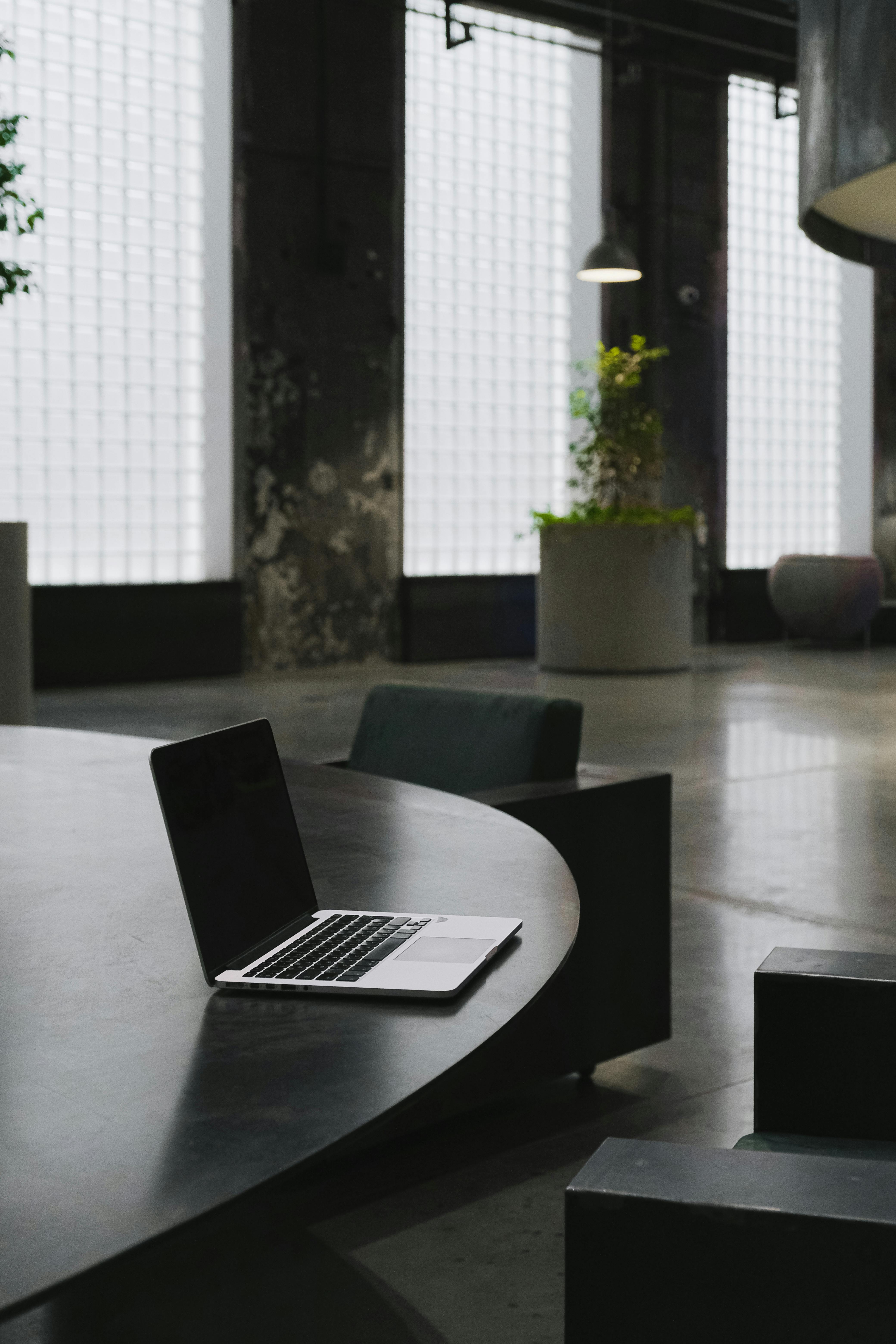 An open laptop sits on a round black table in an industrial-style room with large glass block windows and potted plants.