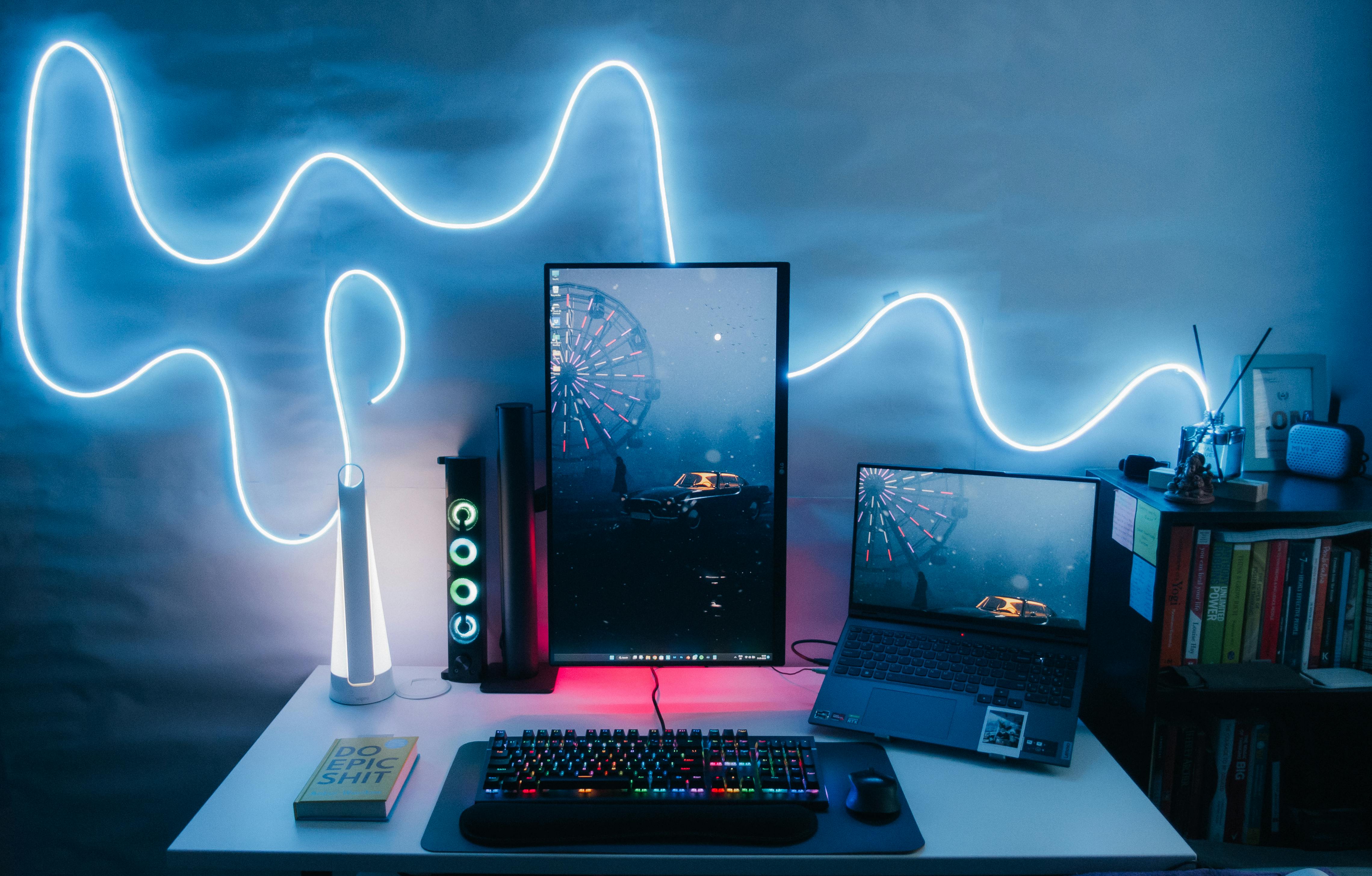 A modern gaming setup includes a vertical monitor and laptop with matching wallpapers, RGB keyboard, mouse, and glowing neon wall lights, creating a cool ambiance.