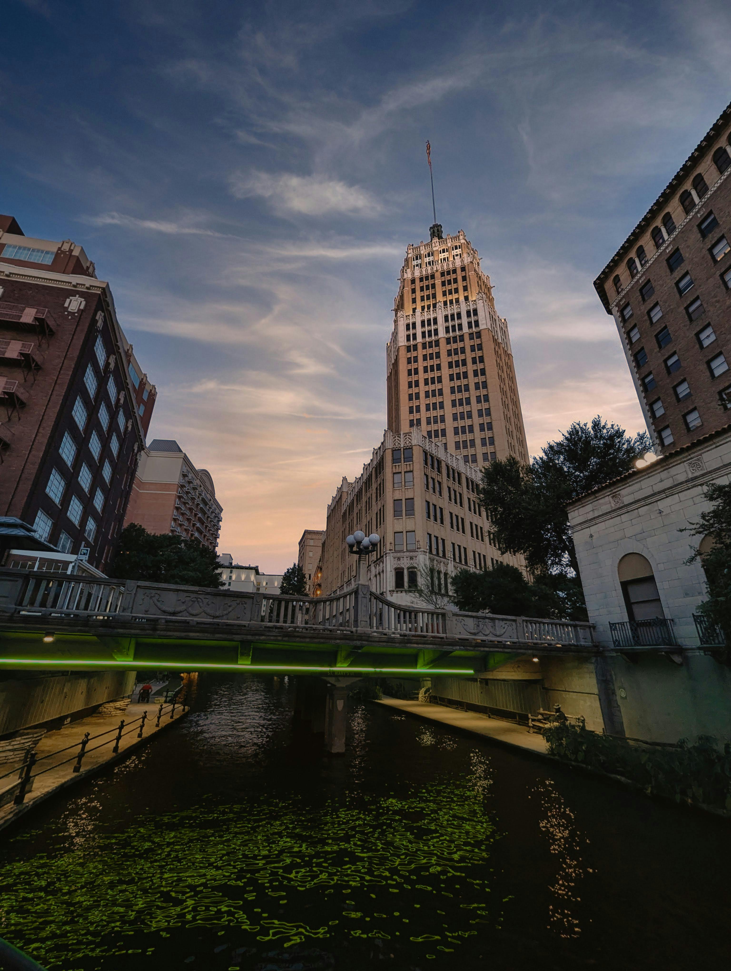View of a tall historic building with an American flag on top, a bridge over a river, and surrounding buildings at dusk.