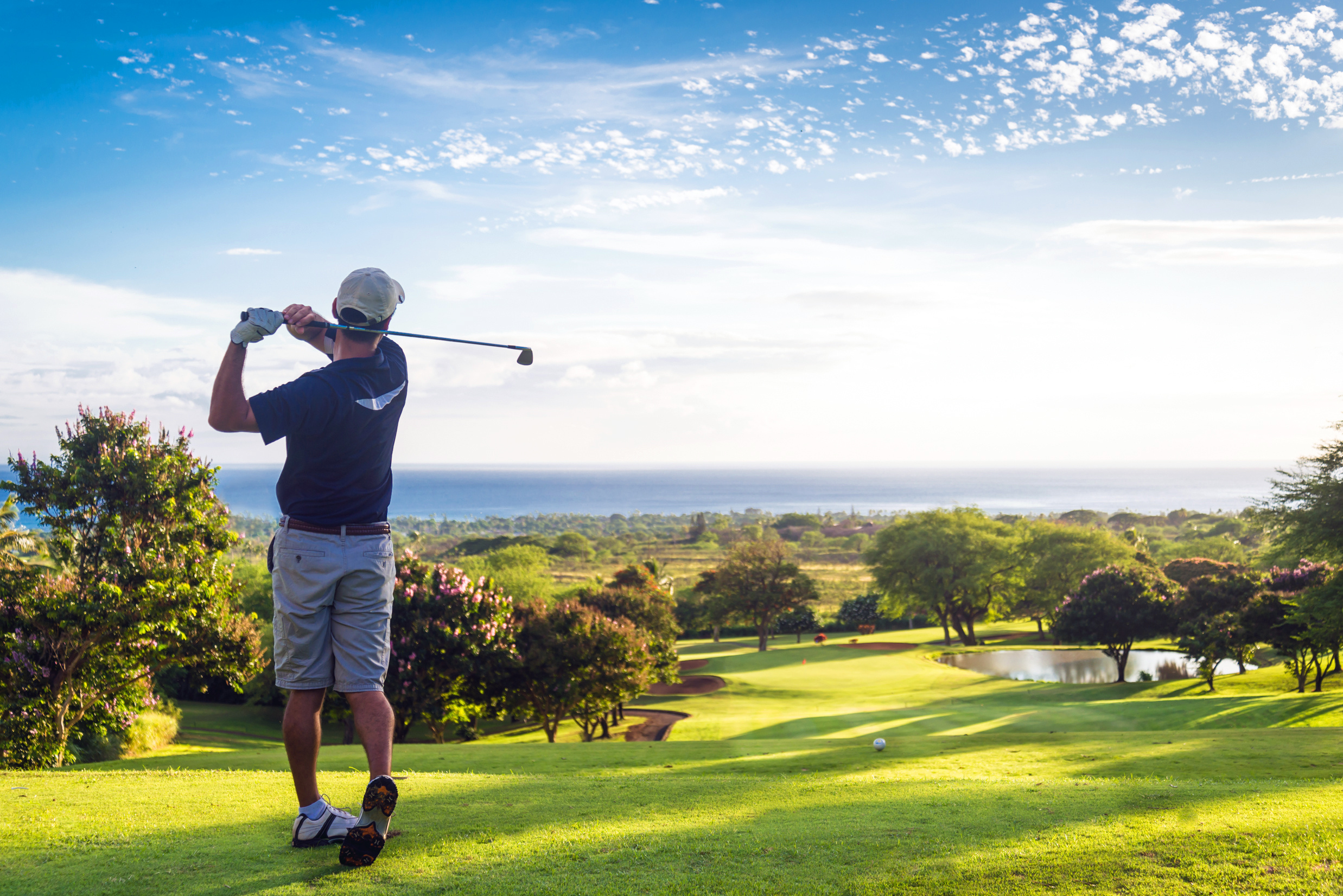 Golfer wearing a cap and gloves swinging a club on a green golf course overlooking trees and water under a partly cloudy sky.