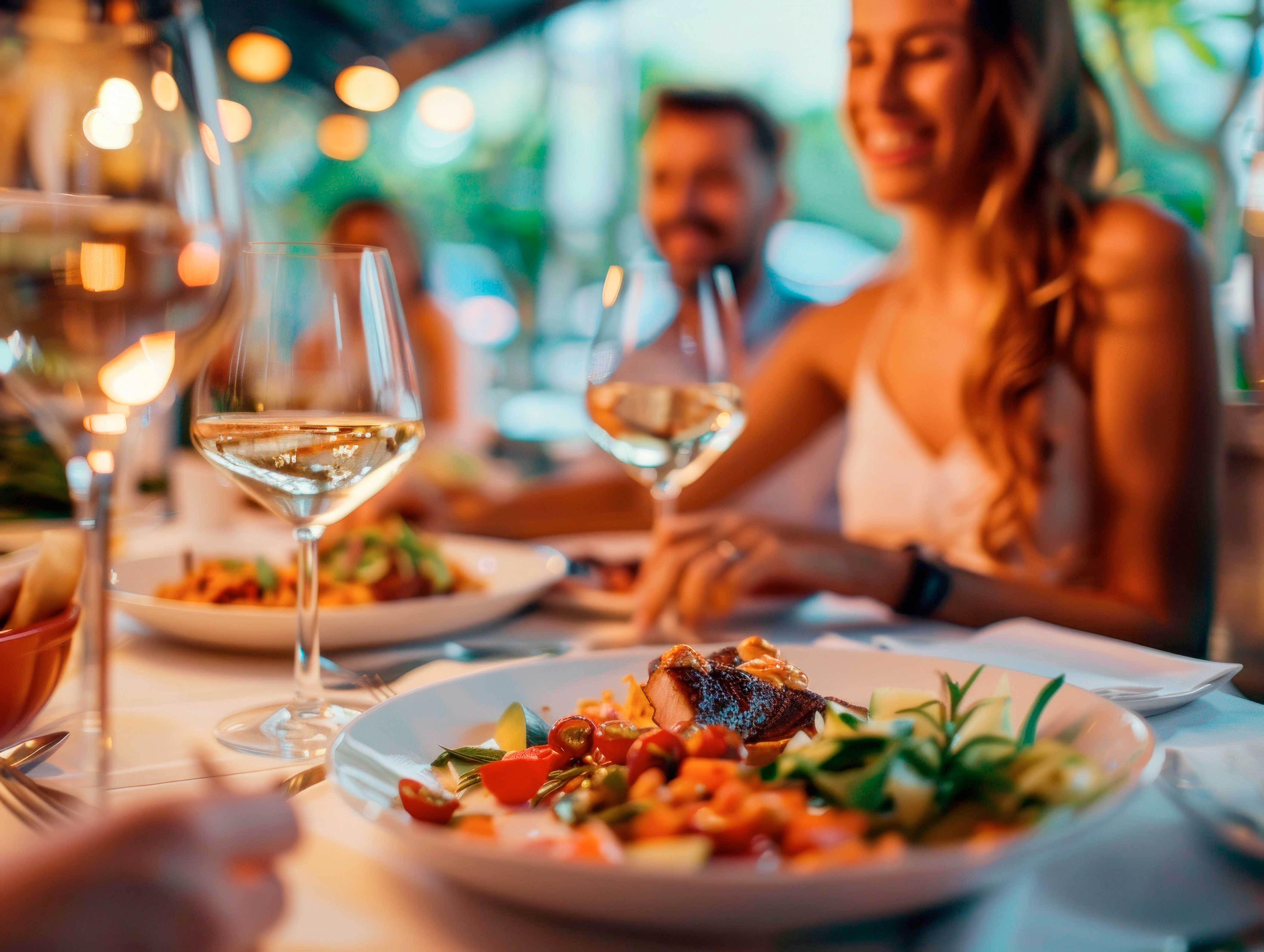 Close-up of a gourmet meal and wine glasses on a table with smiling people dining in the background.