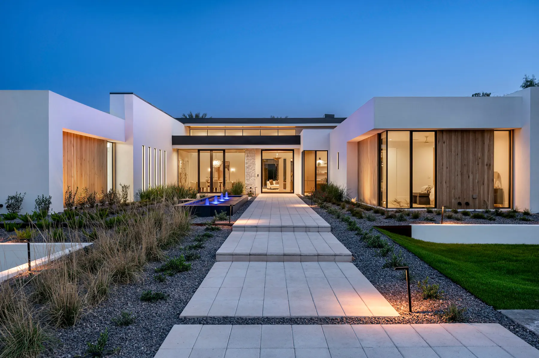 Modern white house with large glass windows, wooden accents, a tiled pathway, and a blue-lit water feature at dusk.