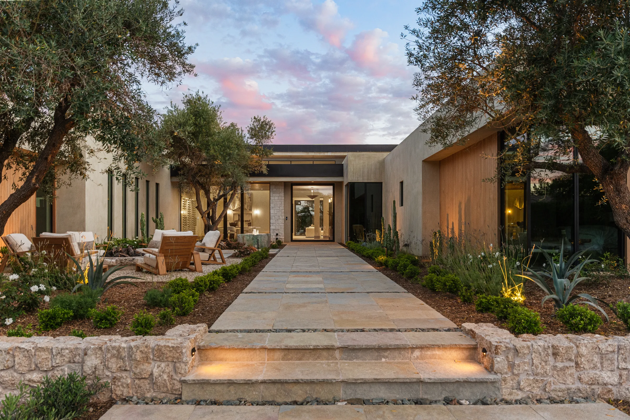 Modern single-story white house with large glass doors, wooden accents, illuminated pathway, and a blue fountain at dusk.