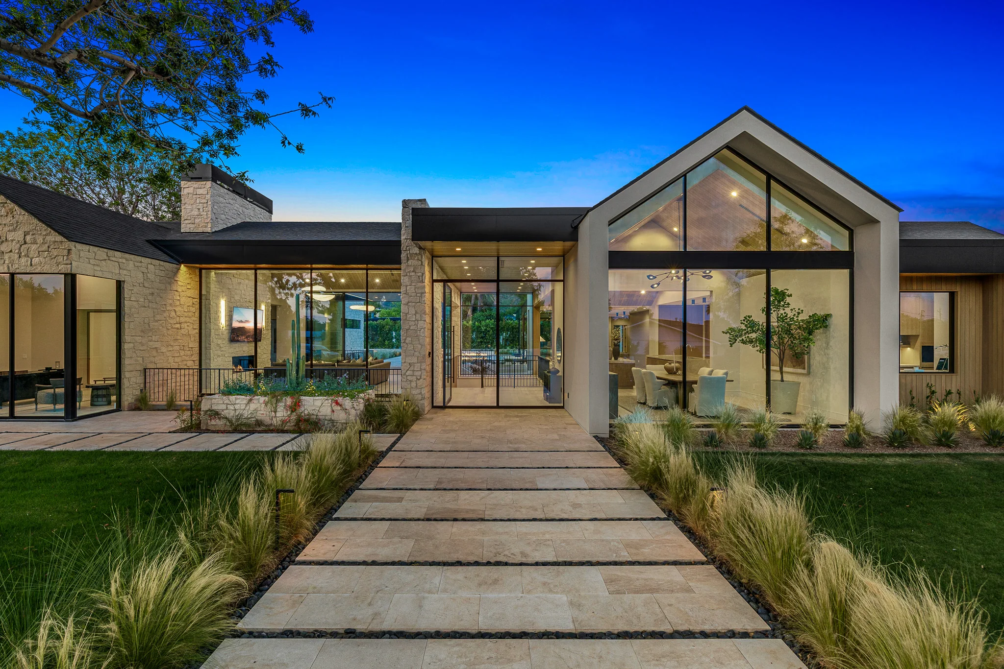 Modern single-story house with large glass windows, stone and wood exterior, surrounded by green lawn and trees at dusk.