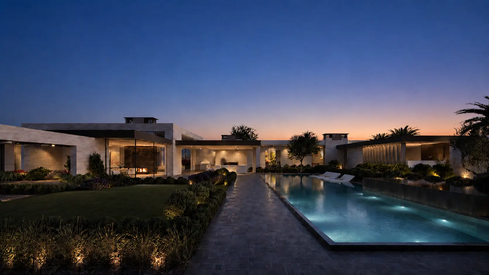 Modern luxury house with large glass windows, an infinity pool with water cascading over stone wall, lush landscaping, and mountain backdrop at dusk.
