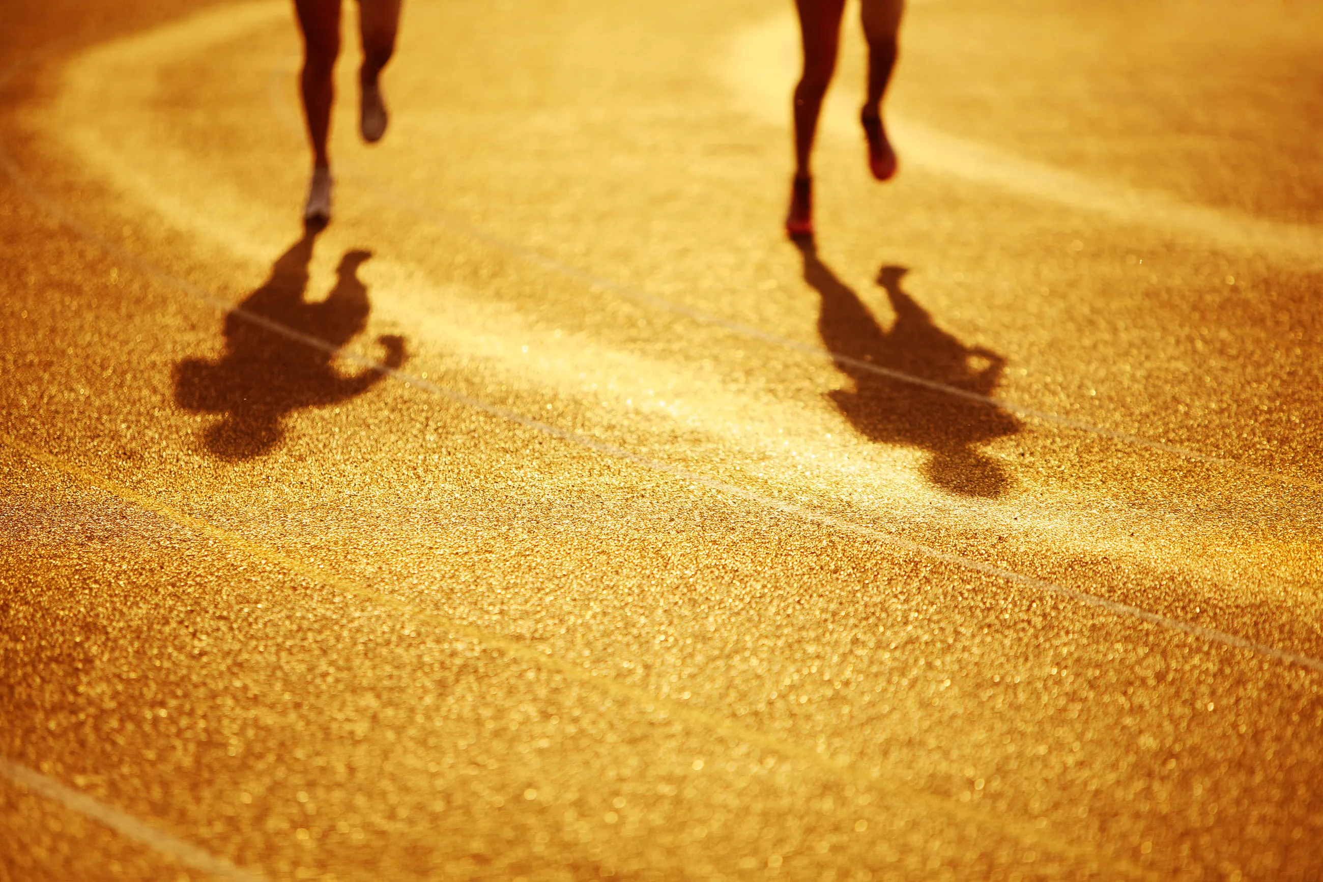 The legs and shadows of 2 athletes running around a track