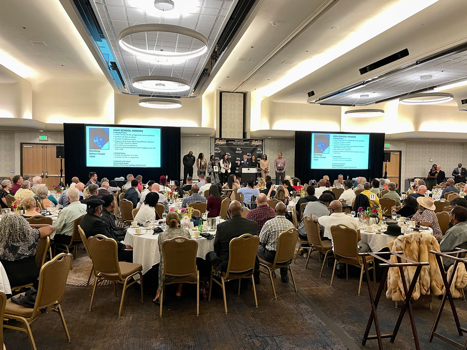 Large indoor banquet with many seated guests facing a stage where six people stand during an awards ceremony with two projection screens showing high school honors.
