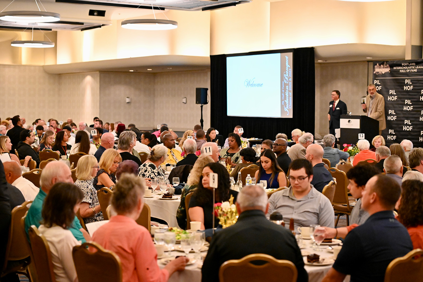 Large banquet hall with many people seated at round tables, listening to a speaker at a podium beside a projection screen displaying a welcome message.