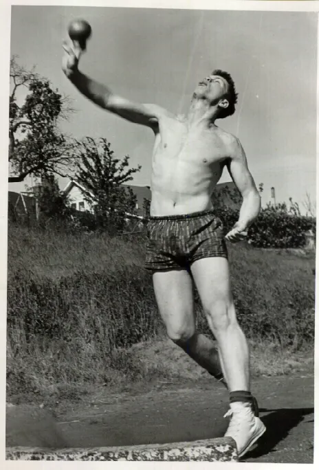 Black and white photo of a shirtless man in shorts throwing a ball outdoors on a sunny day.