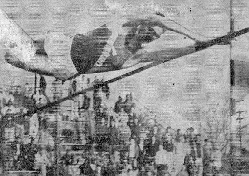 Black-and-white photo of an athlete mid-air performing a pole vault over the bar with a crowd watching in the background.