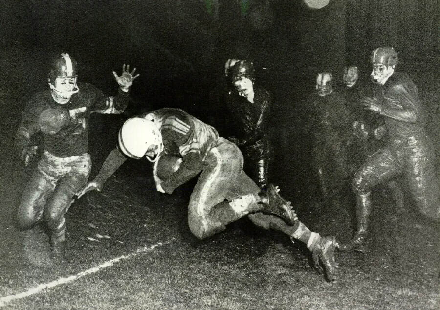 Black and white photo of a vintage football game with a player in a white helmet running with the ball while others in dark helmets try to tackle him.