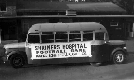 Black and white photo of a bus with a banner advertising a Shriners Hospital football game on August 13th.