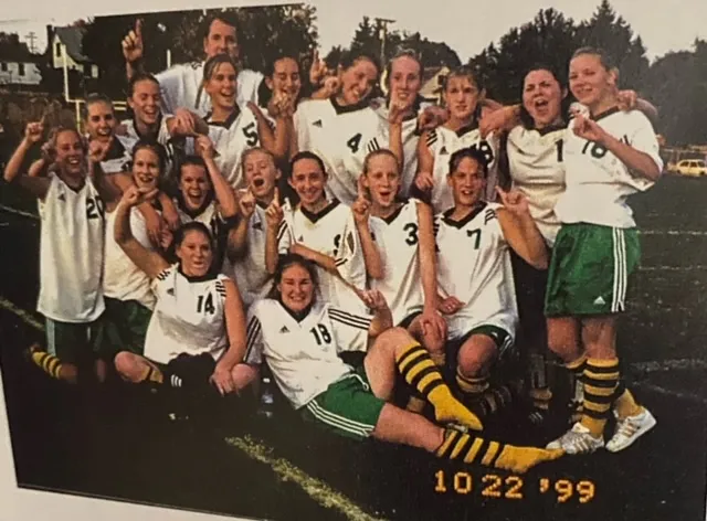 A girls' soccer team posing together on a field, many holding up one finger, wearing white jerseys and striped yellow and black socks, dated October 22, 1999.