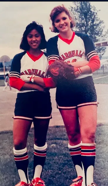 Two female softball players in Marshall uniforms with striped socks standing side by side on a field, one holding a glove.