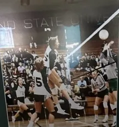 Indoor volleyball game with players from two teams jumping and reaching for the ball near the net in a gymnasium filled with spectators.