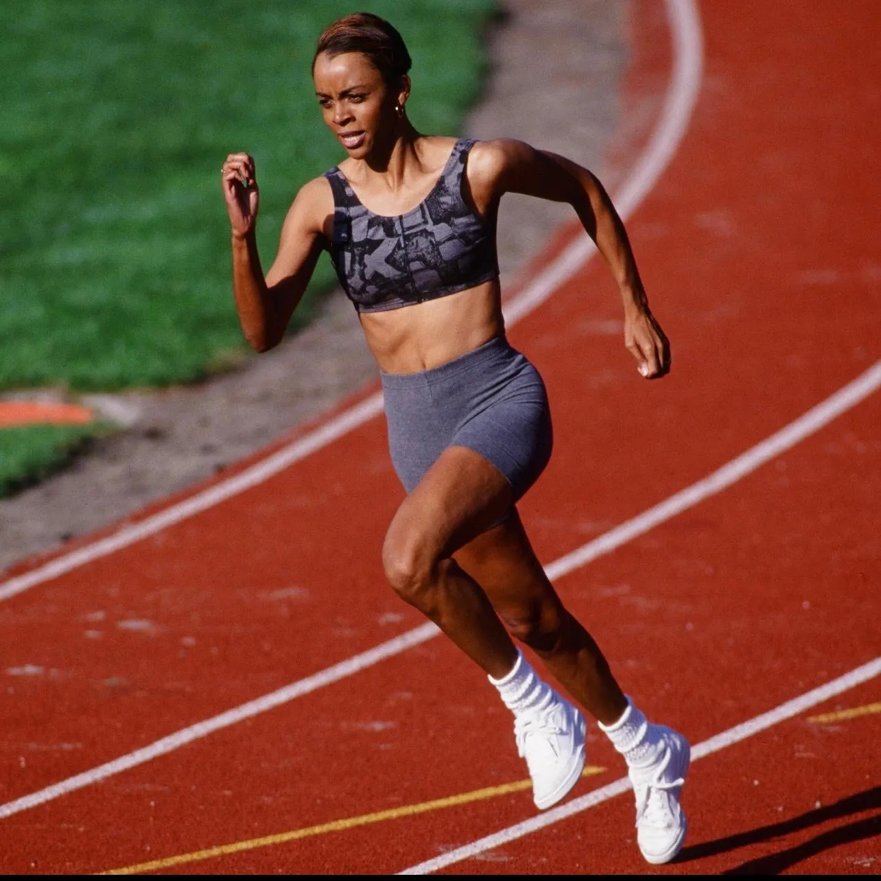 Female athlete sprinting on a red running track wearing a sports bra, shorts, white socks, and white running shoes.
