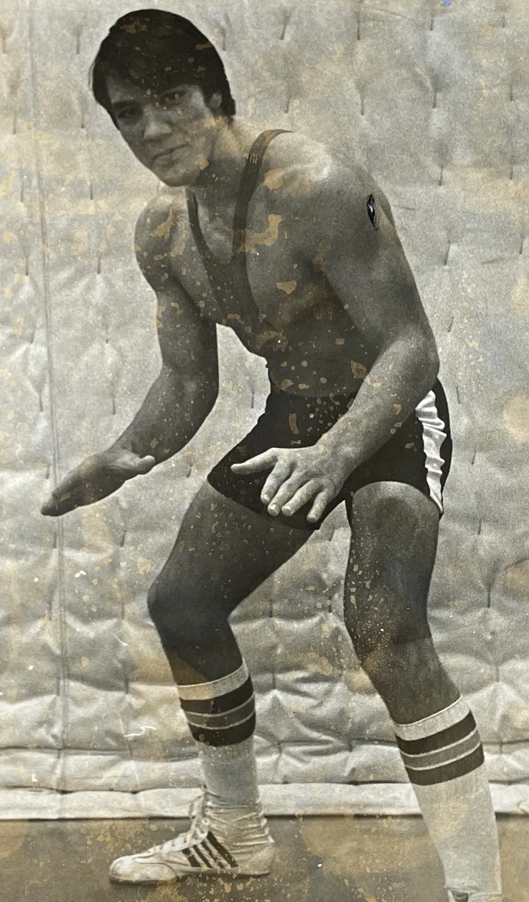 Vintage black and white photo of a wrestler in a crouched stance wearing a singlet, knee-high socks with stripes, and wrestling shoes.