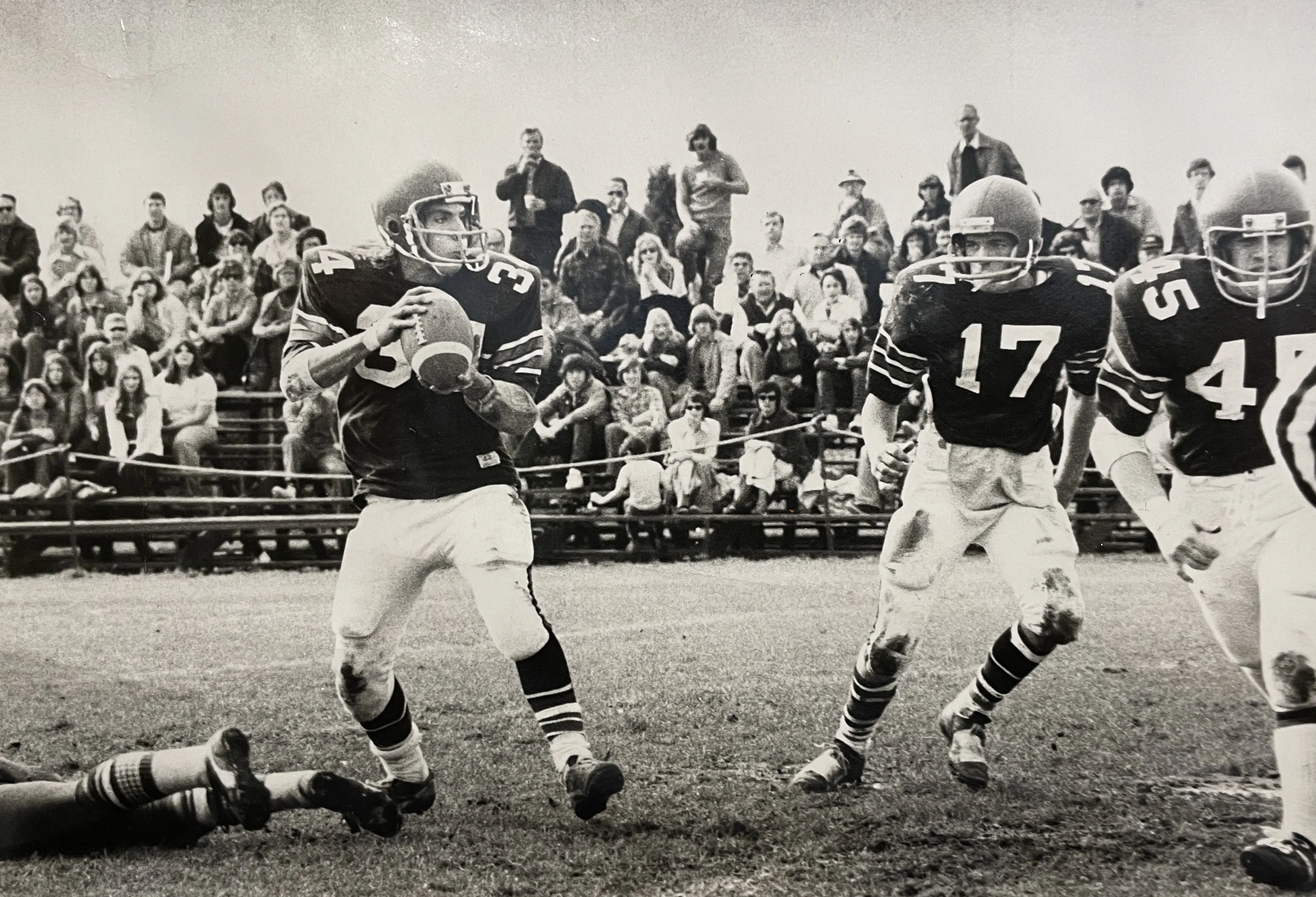Black and white photo of a football game with a player in jersey 34 holding the ball, preparing to pass, while players 17 and 45 move beside him and a crowd watches in the background.