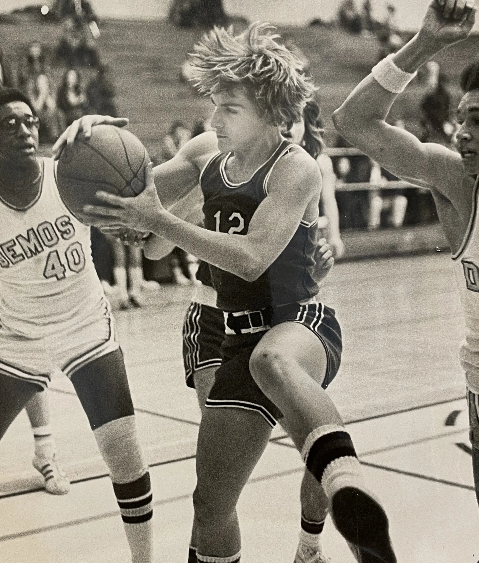 Black-and-white photo of a basketball player in a dark jersey number 12 jumping and holding the ball while defended by two players in white jerseys.