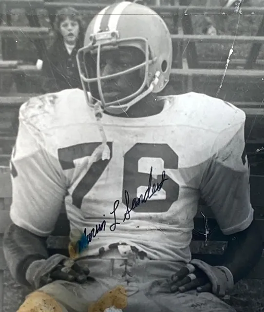 Black and white photo of a football player wearing helmet and jersey number 76, sitting on a bench with a signed autograph on his jersey.