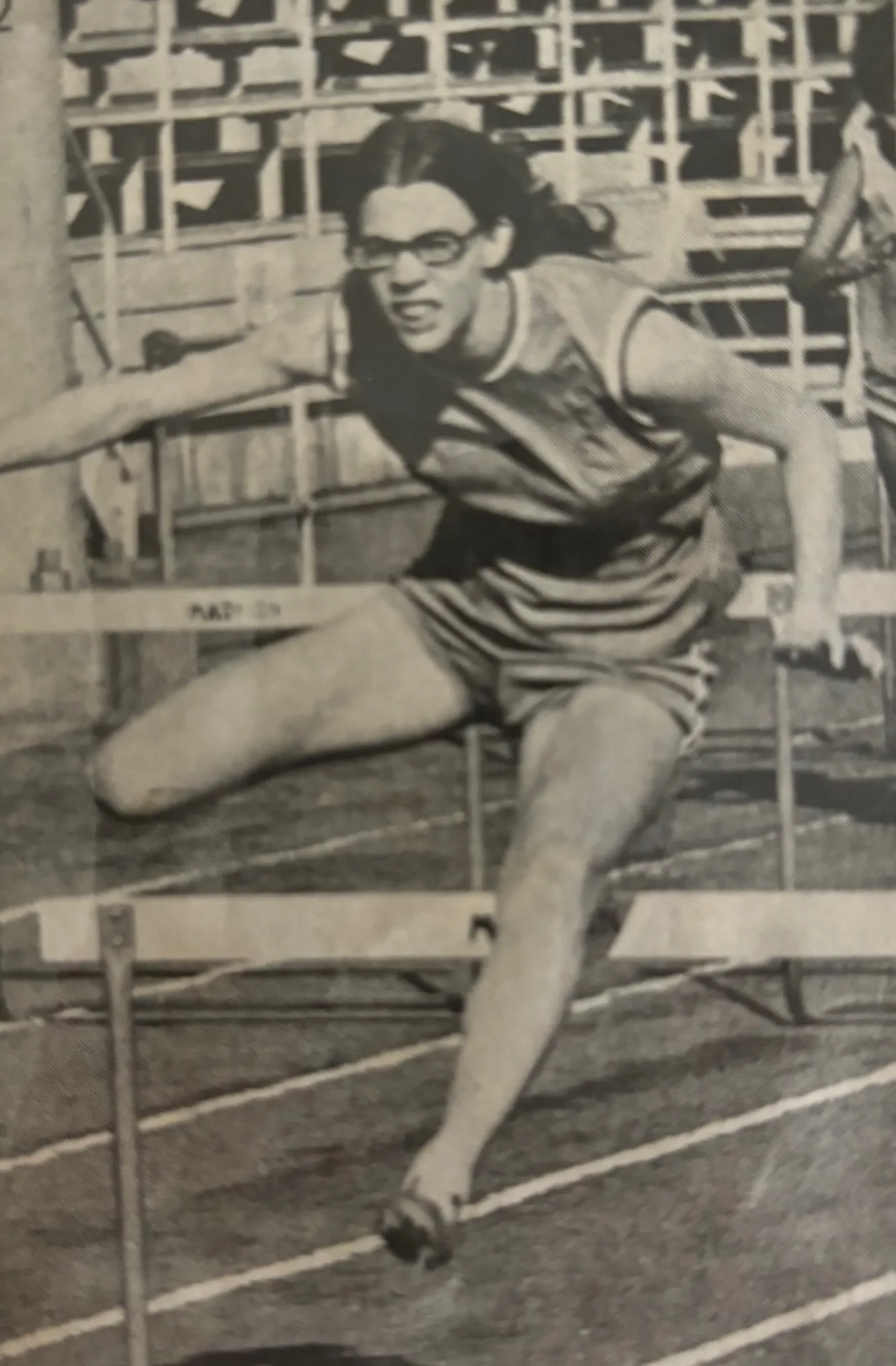 Athlete with glasses mid-jump over a hurdle during a track and field race.