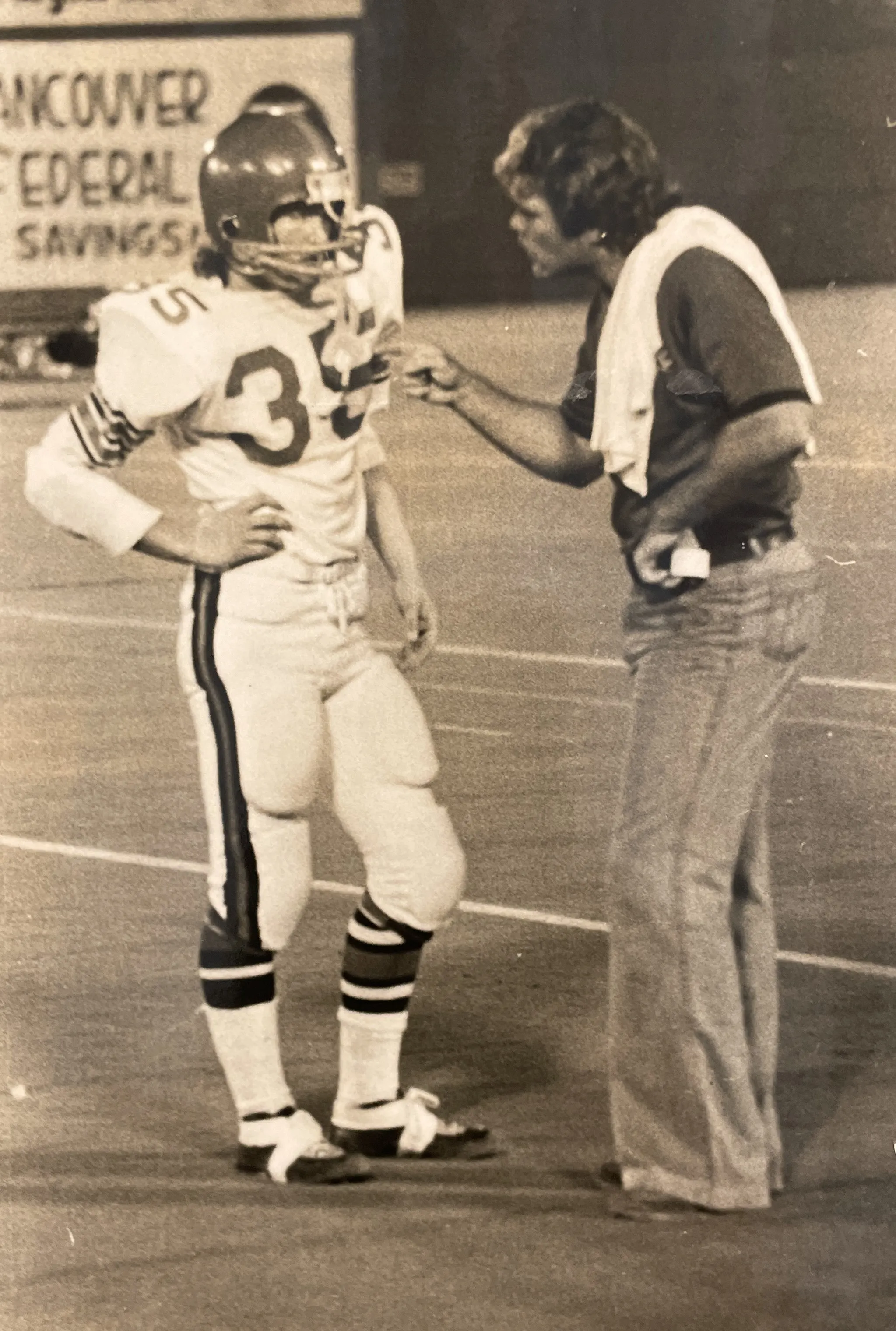 Black and white photo of a football player in uniform and helmet listening to a coach who is speaking and gesturing emphatically on the field.