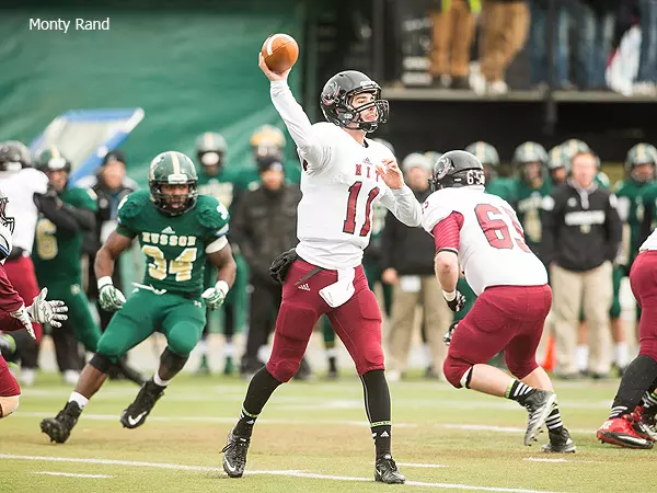 Football quarterback in white jersey throwing a pass during a game, with a defender in green approaching.