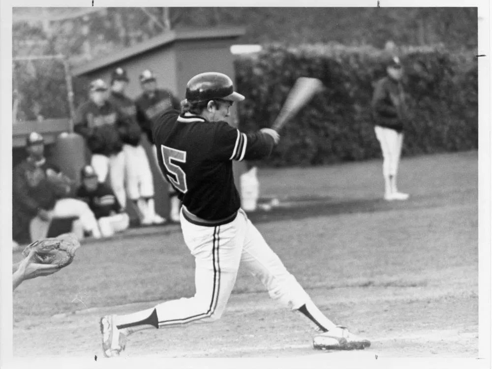 Baseball player wearing number 5 swinging a bat during a game, with teammates and coaches standing in the background.