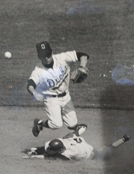 Black and white photo of a baseball player in a Ducks uniform jumping over a sliding player during a game.