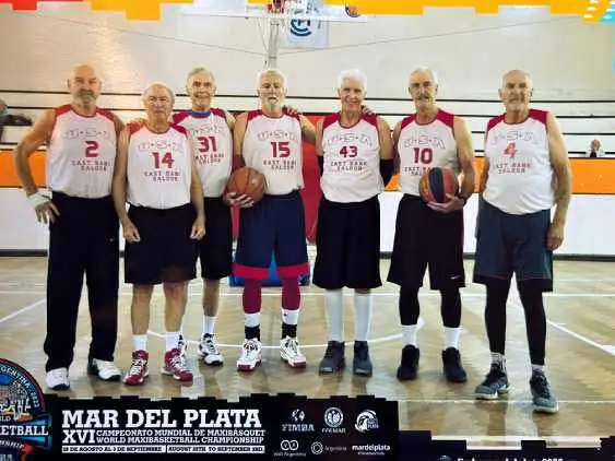 Seven elderly men standing in a basketball gym, wearing matching maroon and white basketball jerseys with numbers, two holding basketballs.