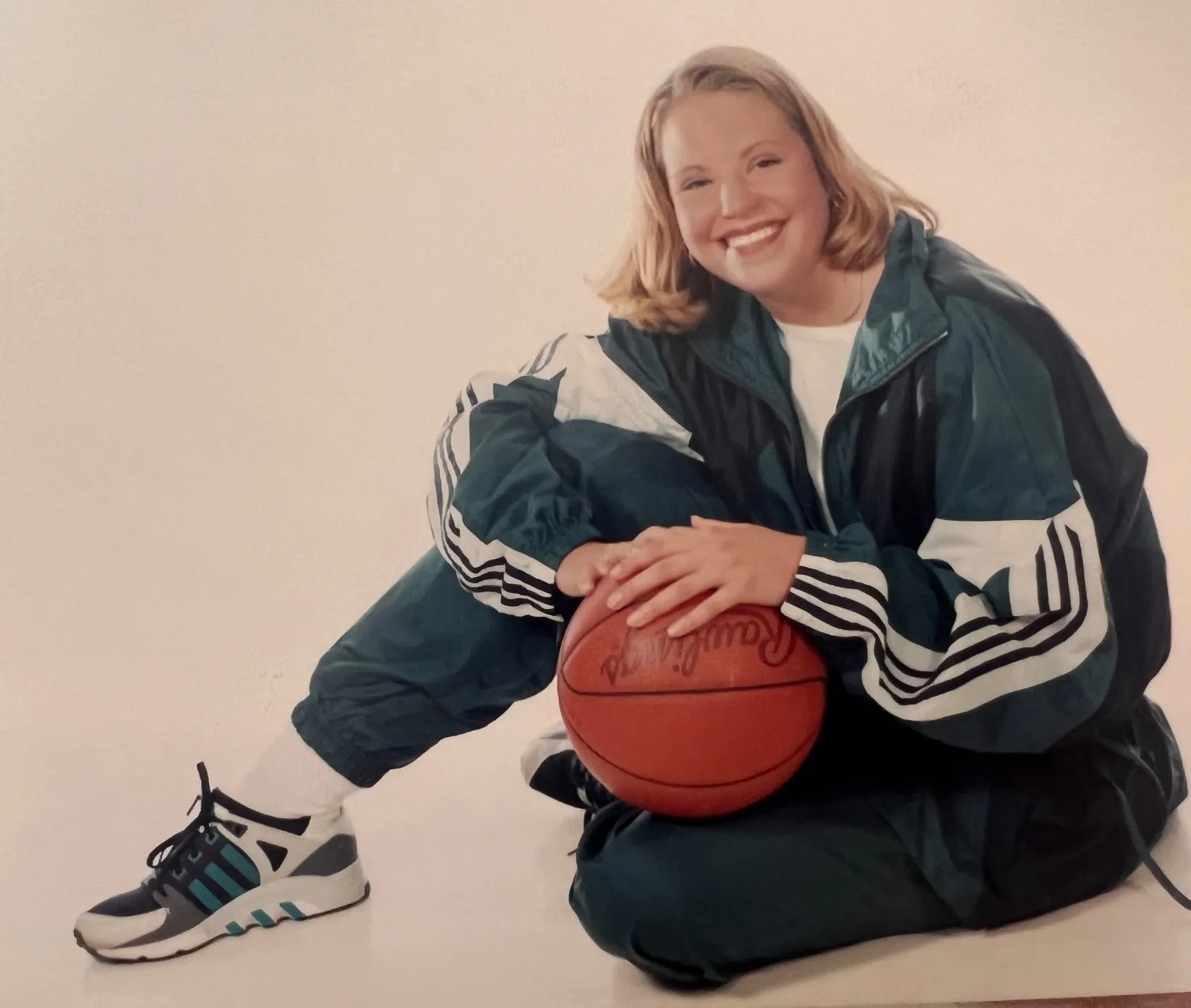 Smiling young woman in a green and white Adidas tracksuit sitting on the floor holding a basketball.
