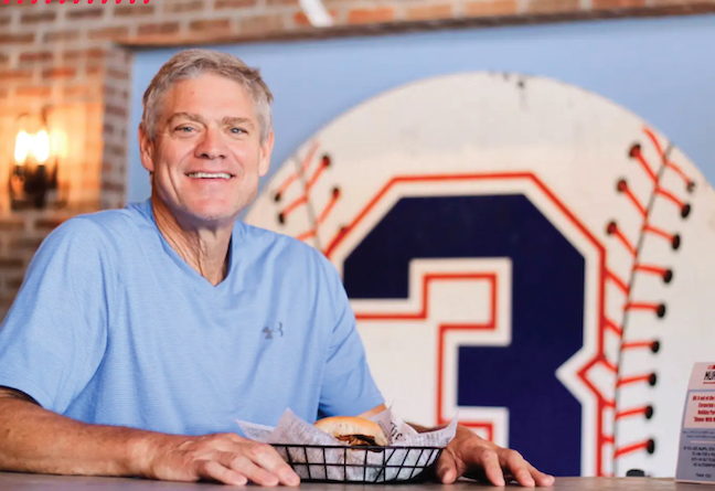 Smiling man in a light blue shirt sitting at a table with a basket of food, with a large baseball-themed decoration featuring the number 3 in the background.