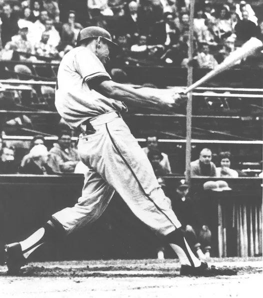 Black and white photo of a baseball player mid-swing at bat in a stadium with spectators in the background.