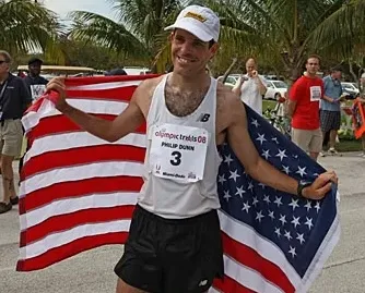 Runner Philip Dunn holding and wearing the American flag after a race, smiling with a white cap and race bib number 3.