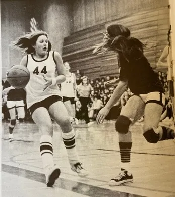 Black and white photo of two female basketball players in action on an indoor court, with one player dribbling and the other defending.