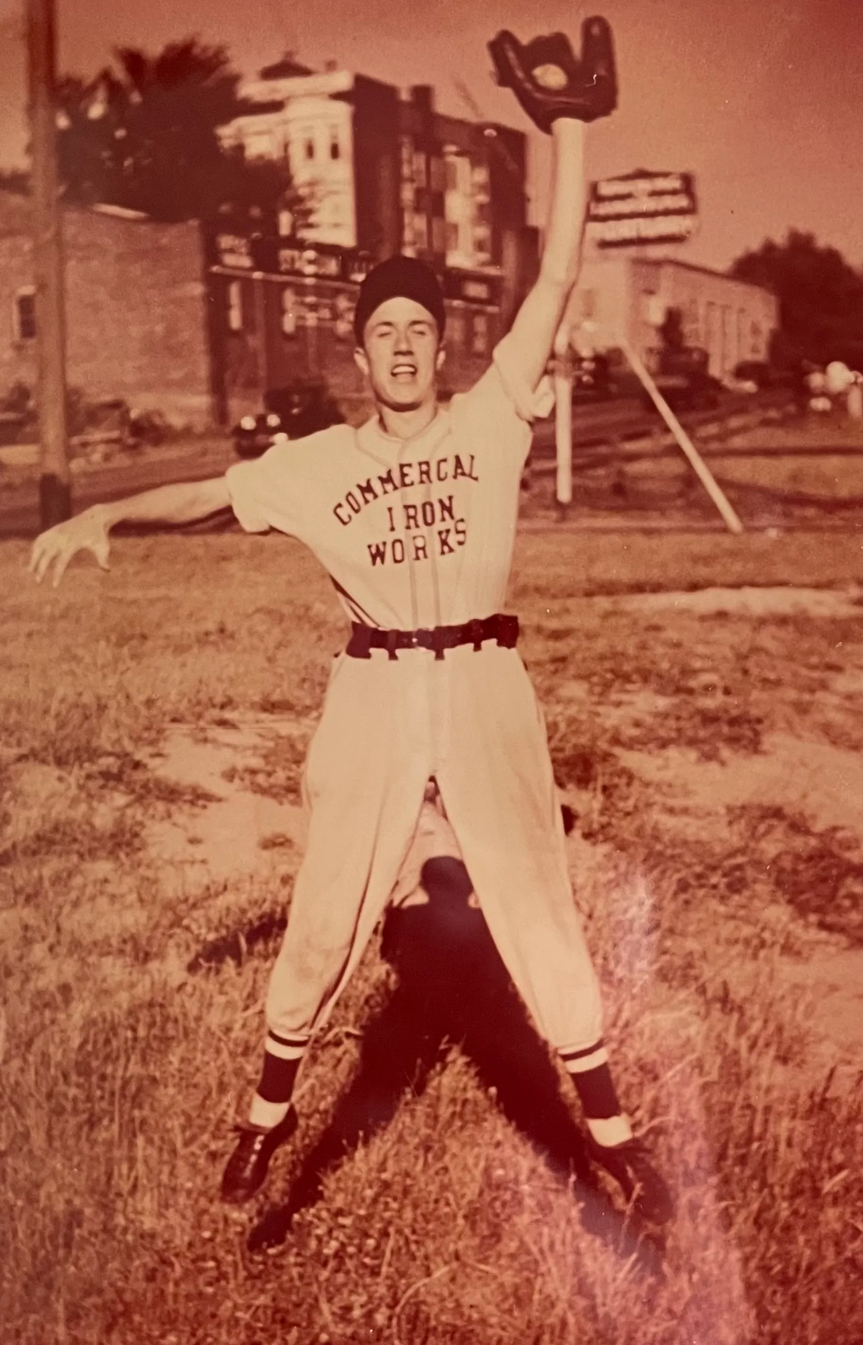 Young baseball player in vintage uniform jumping with glove raised in the air on a grassy field with buildings in the background.