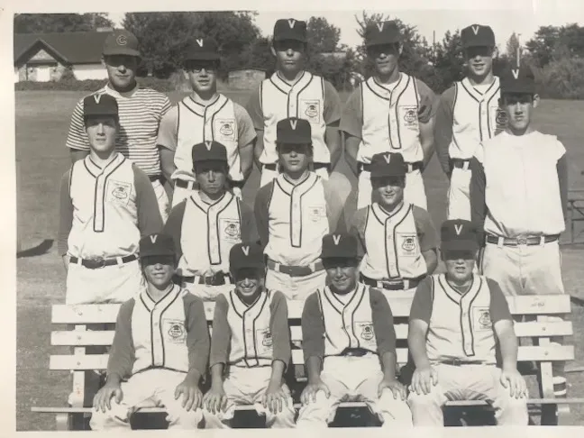 Black and white photo of a youth baseball team wearing 'V' hats and striped jerseys, posing outdoors on a bench with two coaches standing behind.
