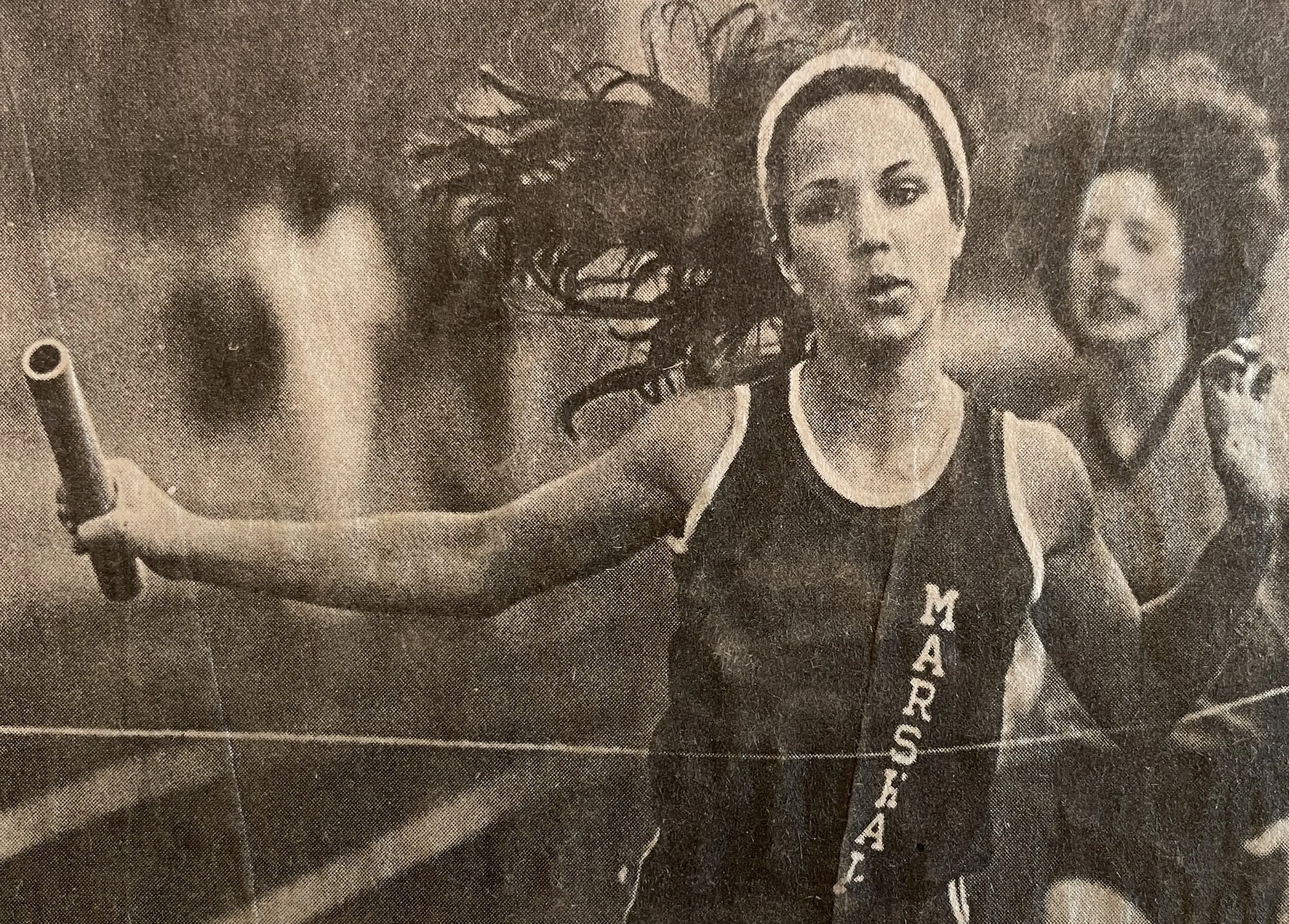 Female athlete crossing the finish line with a baton in hand wearing a Marshals track uniform during a relay race.