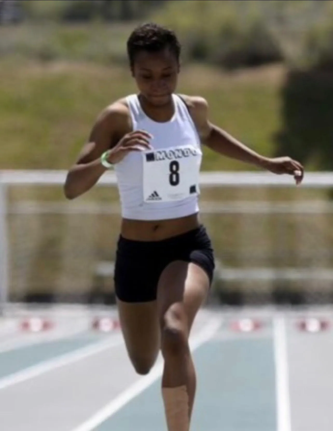 Female athlete in a white tank top and black shorts sprinting on an outdoor track during a race.