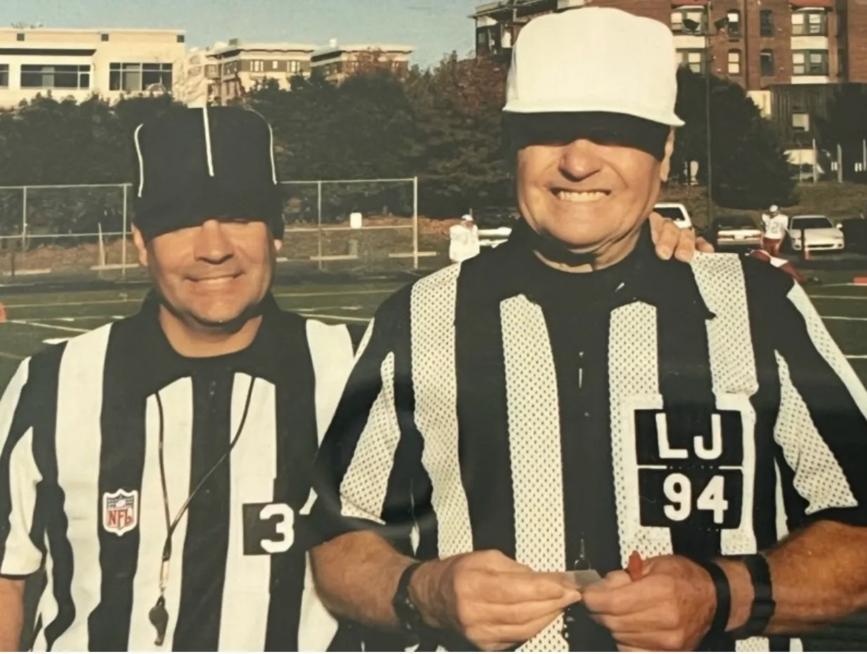 Two football referees in black-and-white striped shirts and hats smiling on a sunny field.