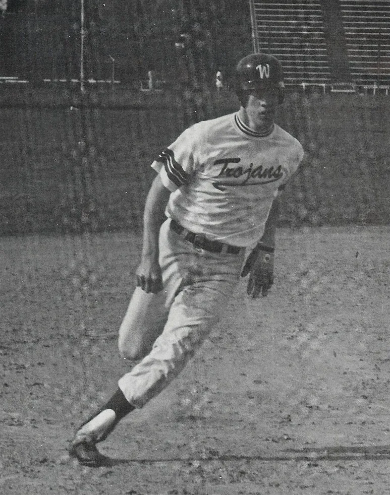 Black and white photo of a baseball player wearing a 'Trojans' jersey and helmet running on a dirt field.