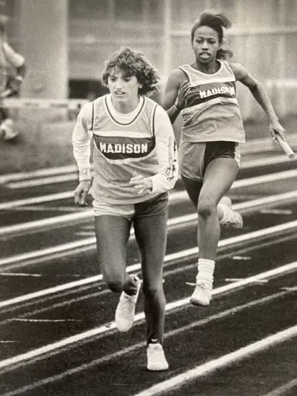 Two female athletes from Madison running a relay race on an outdoor track, one holding a baton.