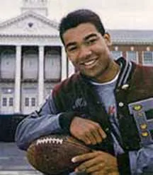 Young man smiling while holding a football in front of a large building with columns.